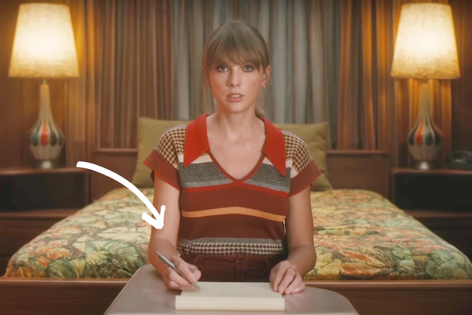 Swift sits a schooldesk in front of a bed in her bedroom, facing the camera. Below her, a notebook, and she holds a pen in her right hand, and, how do I describe this, it's sort of a claw grip, with her pen nestled not between her thumb and index finger, as is normal, but instead its entire length is between her index finger and her middle finger.