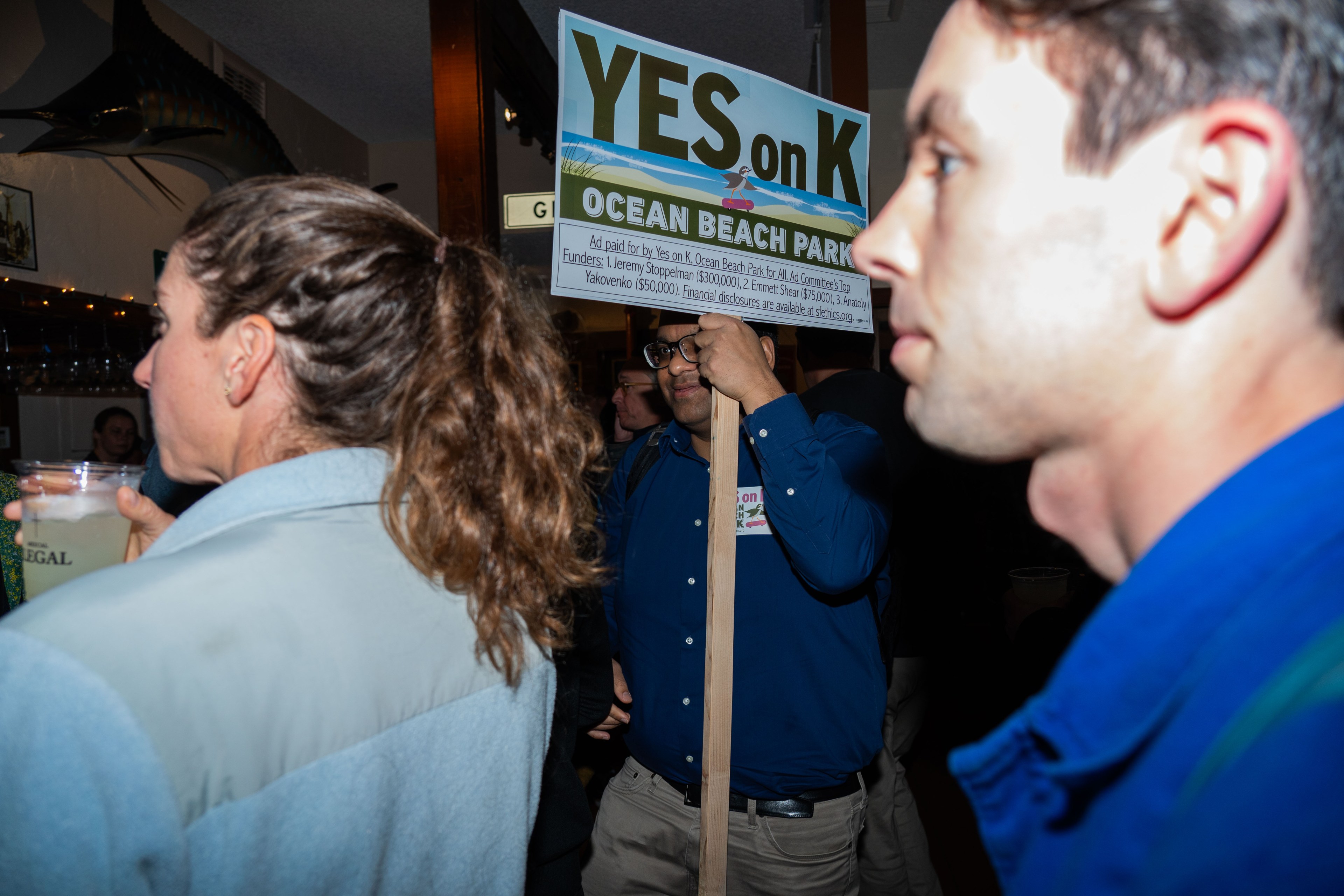 The image shows people at a gathering. A person holds a sign advocating for "YES on K" regarding Ocean Beach Park. Others nearby are engaged in conversation.