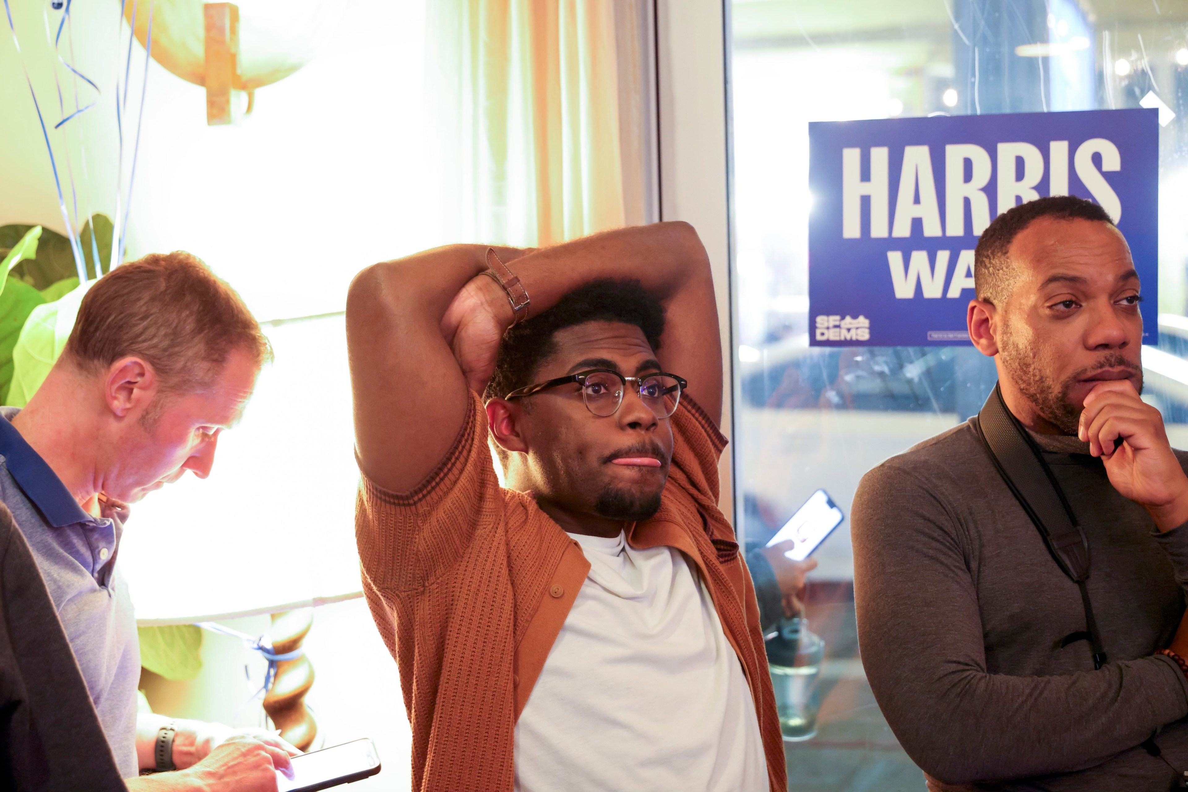 Three men are indoors, one checking his phone, another with hands on his head, and the third looking thoughtfully. A "Harris" sign is visible behind them.