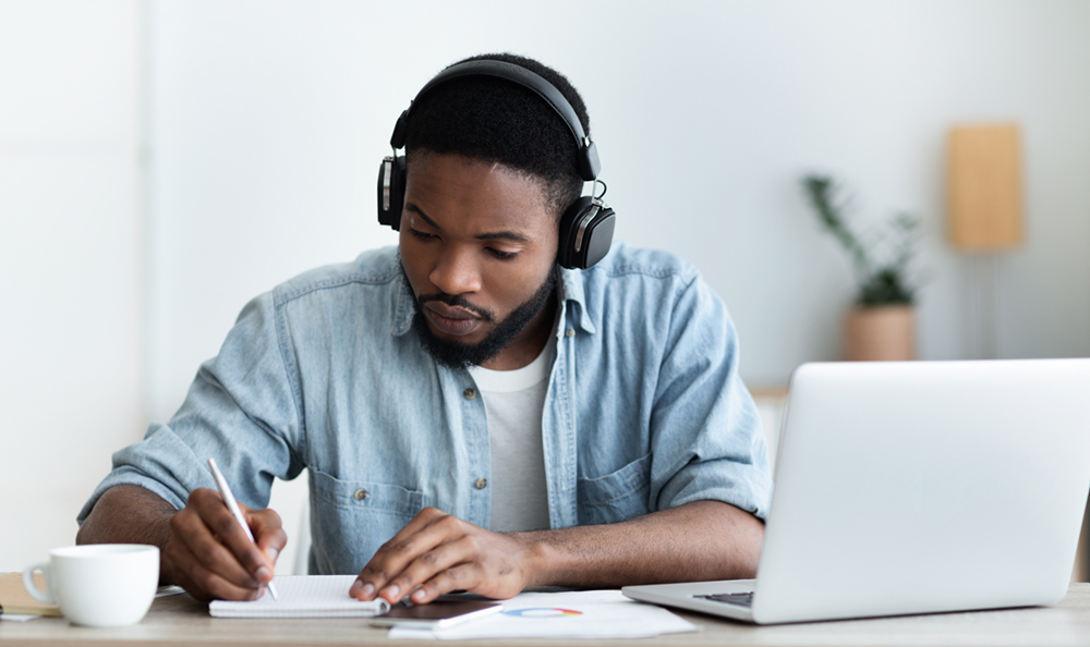 jeune homme noir portant des écouteurs et écrivant des notes assis à un bureau avec un ordinateur portable