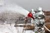 Firefighters in protective silver suits spray a thick white foam from a large hose