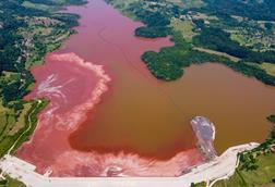 A lake from above with red sludge spreading across its brown waters. There are houses and trees on the banks.