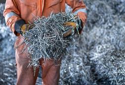 A person in overalls and protective gloves holding a mass of curled metal strips next to a large pile of the same metal strips