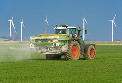 A tractor spreading fertilizer pellets on a field with wind turbines in the distance