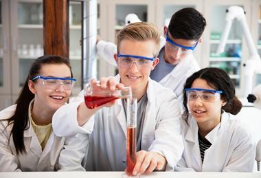 Four high school students in a practical chemistry lesson wearing goggles and lab coats