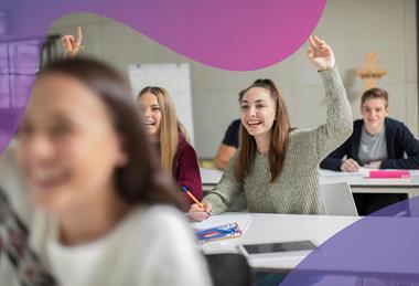 Two female students wearing casual clothes sit at a desk, raising their hands and smiling