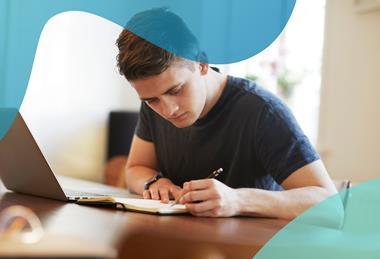 A young man works at a table, writing in a diary with a silver laptop open next to him