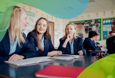 A group of three students wearing school uniform sit at a desk in their classroom talking, with their notebooks open in front of them