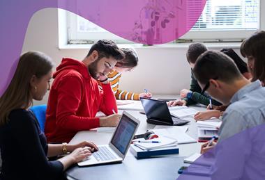 A group of students wearing casual clothes work together at a table using laptops, pens and notebooks