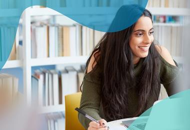 A female student smiles while sitting at a desk in a library, with books on shelves behind her