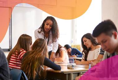 A female teacher talks with a group of students at a table in their classroom, while two of the students make notes