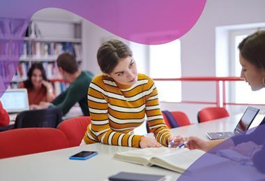 Two students sit opposite each other at a desk in a library, discussing a book open between them and pointing at a paragraph with their pens