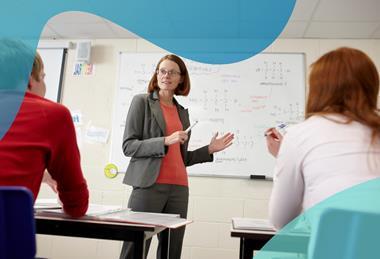 A teacher stands in front of two students sitting at desks; behind her, a whiteboard shows the chemical structures of a variety of compounds