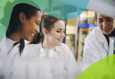 A female teacher and two students wearing white lab coats smile while observing apparatus set up for an experiment