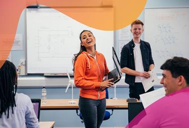 A female student carrying a laptop stands at the front of a class laughing with fellow students, with a model wind turbine and a whiteboard with a circuit diagram behind her