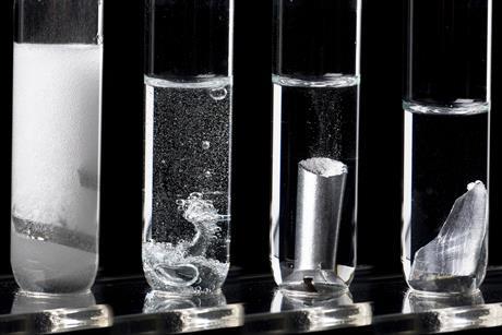 Four test tubes in a row with metals reacting in a clear liquid increasing in bubbles from left to right