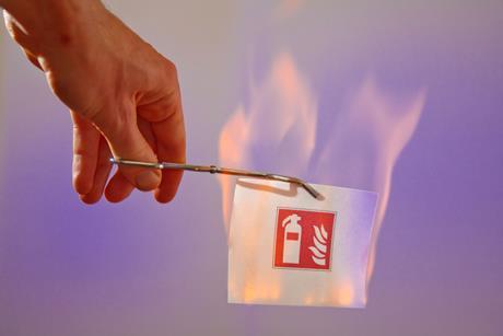 A hand using scissor-handle tweezers to hold a piece of paper that is on fire but not burning