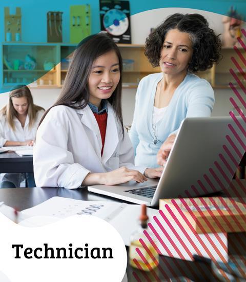 A teacher in a classroom talking to a student wearing a lab coat and using a laptop computer