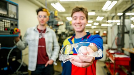 Two students in the background and one is holding safety goggles in the forefront