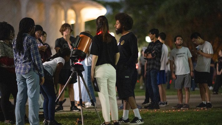 People gather around a telescope on a lawn with Beckman Auditorium in the background