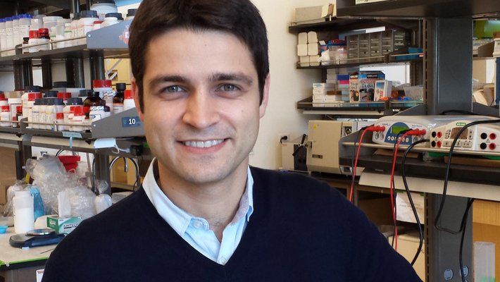 A portrait of Mikhail Shapiro in a laboratory. He wears a sweater and collared shirt. Equipment is seen in the background.