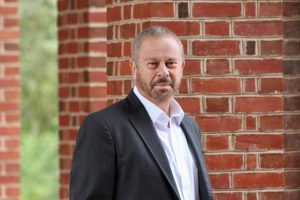 A photograph of Ed Schlesinger standing in front of a brick column