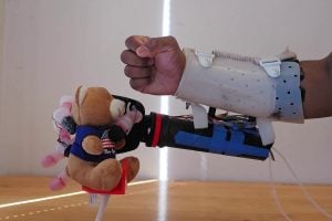 A human arm guides a robotic arm in grasping a teddy bear by mimicking the intended grip.