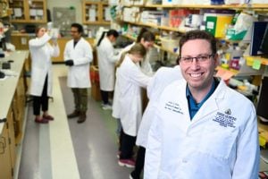 Professor Jordan Greene poses for a photo, while behind him, students and researchers work in a lab.