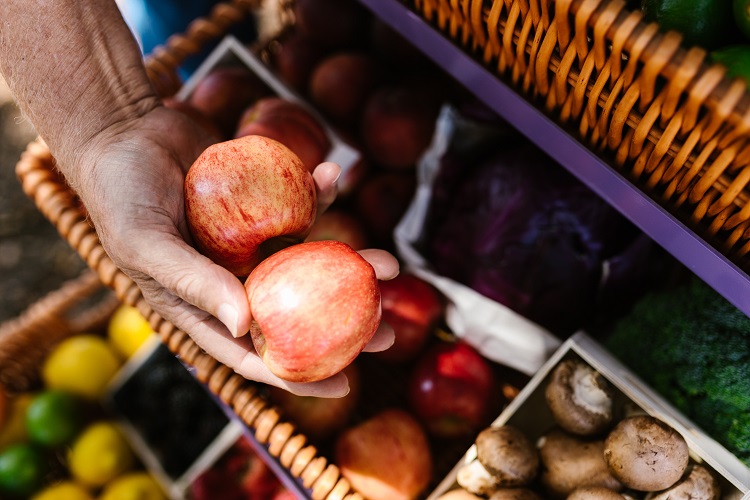 A hand holds two apples over baskets of fruits and vegetables