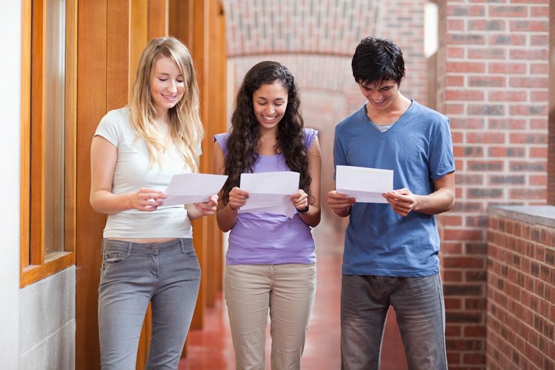 Three high school students, two girls and one boy, look intently at pieces of paper with their exam results on them and smile