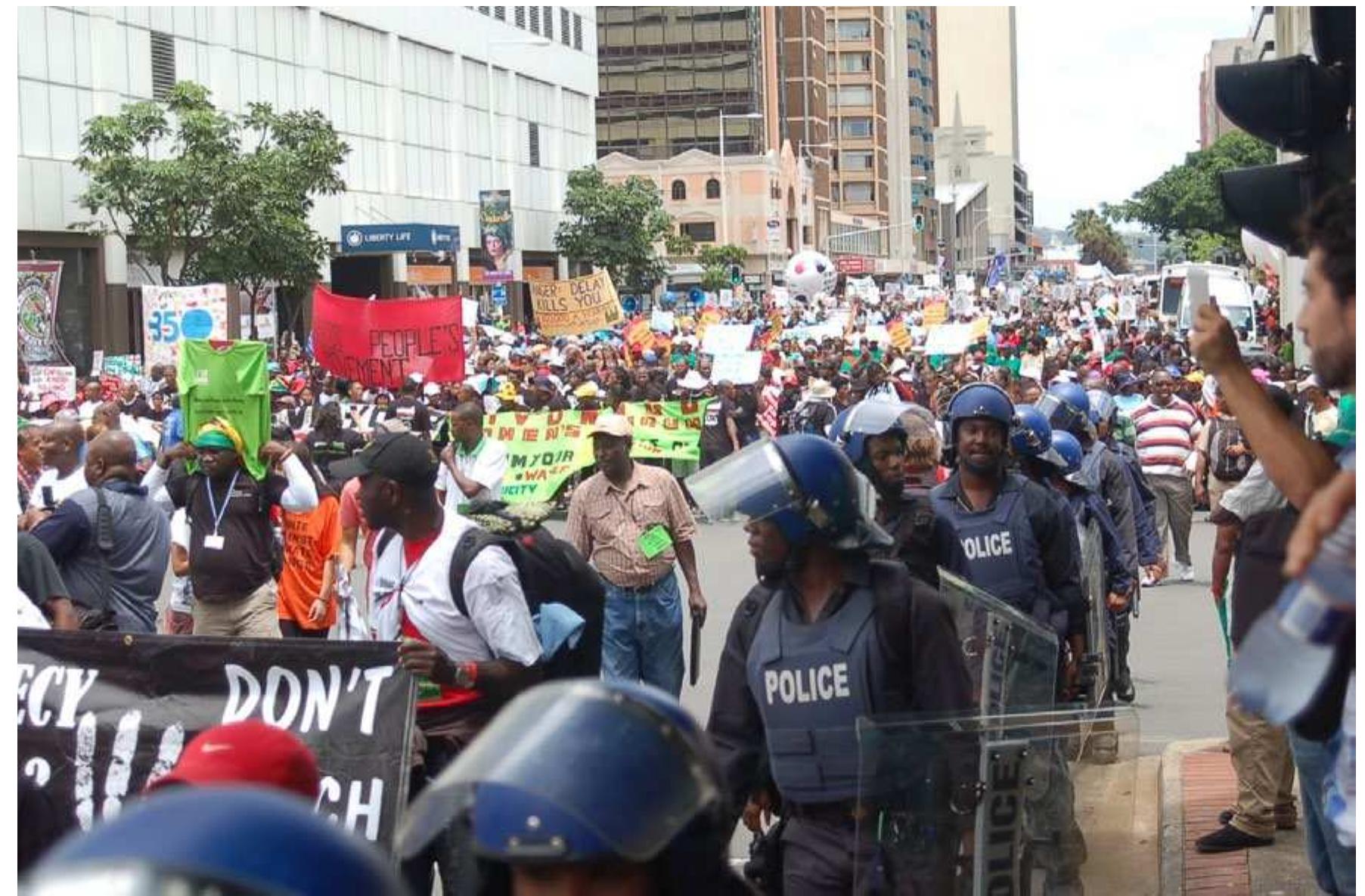 Photo Credit: Tamra Gilbertson, Carbon Trade Watch, Durban, South Africa, 3 December 2011  indictment of our ruling class as an agent of global capital and its economic policies which were deepening inequality and increasing poverty.  Sadly, no matter how hard South Durban Community Environmental Alliance leaders tried to organize in the weeks preceding the COP17 in the city’s most radical anti- corporate protest site (where I too am a lay-member and resident), Africa’s industrial armpit could not consistently deliver more than a few hundred protesters from the 300,000 victims residing in the vicinity. 