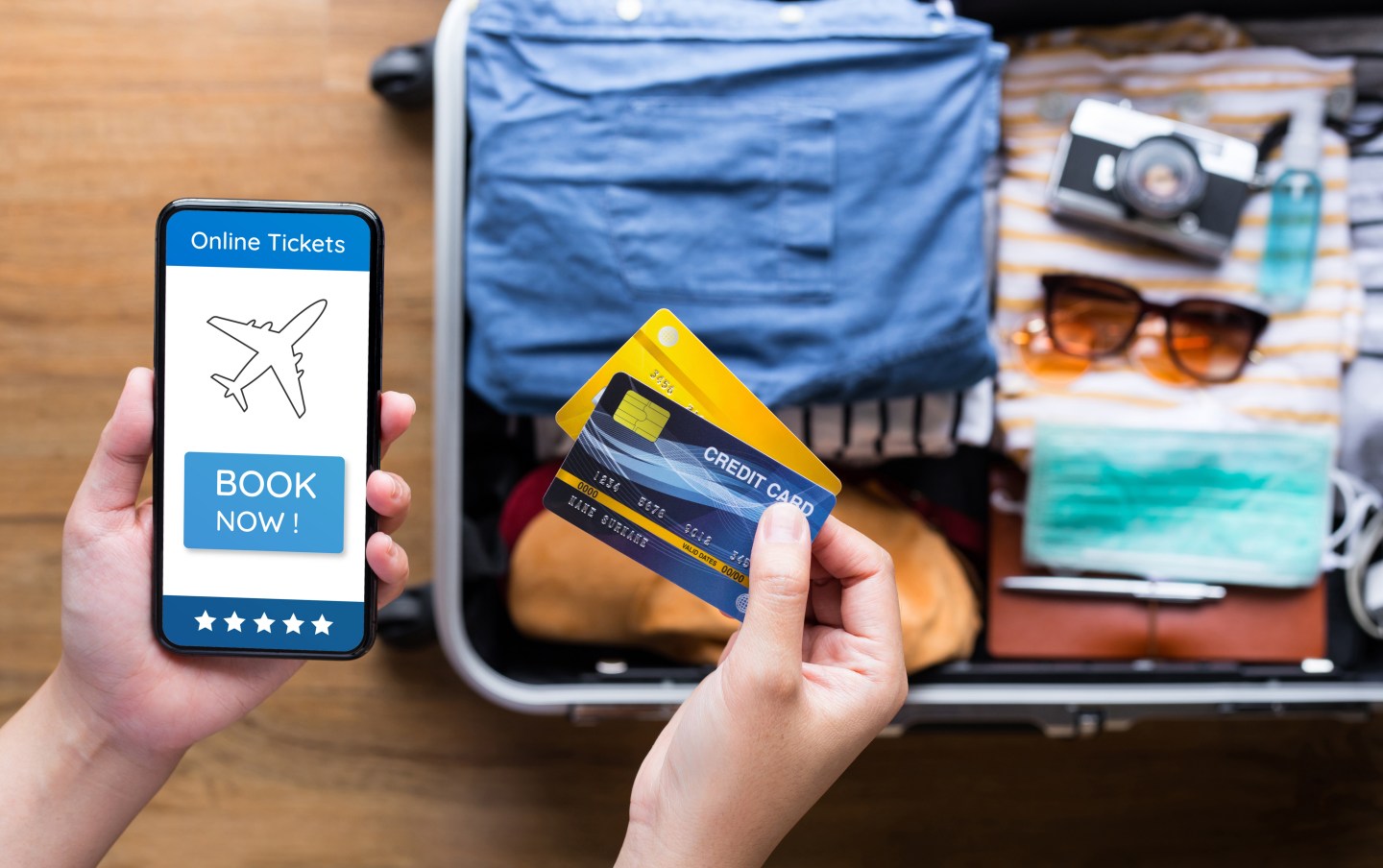 Photo of an open packed suitcase with a person standing over it using credit cards with smartphone for payment of a ticket or booking hotel.