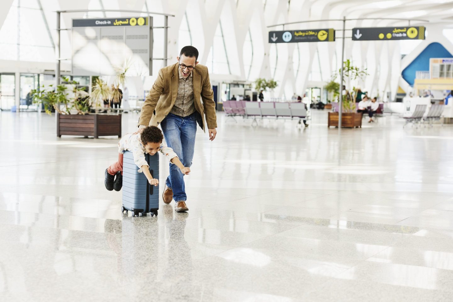 Wide shot of father pushing son riding on rolling luggage through airport terminal.