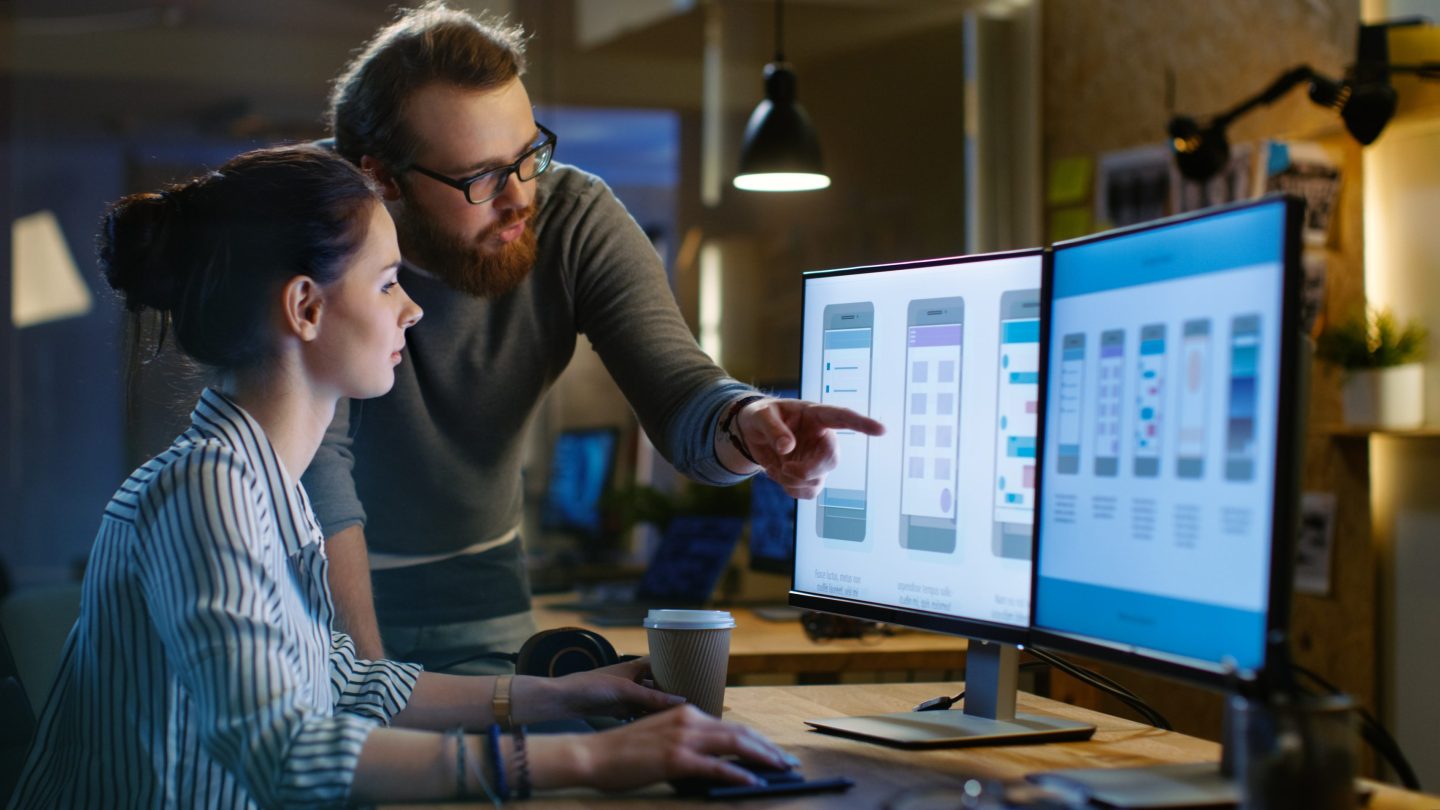 Female UX designer has discussion with male design engineer. They work on mobile application late at night; she drinks coffee.