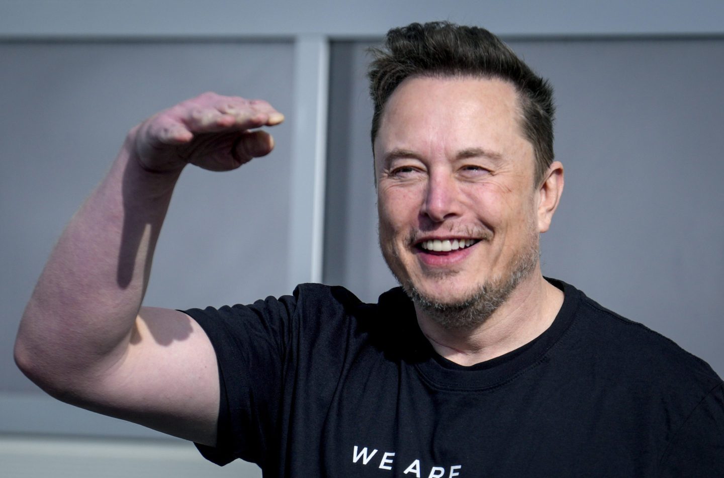 Elon Musk is wearing a black shirt and smiling and he lifts his hand up to his face.