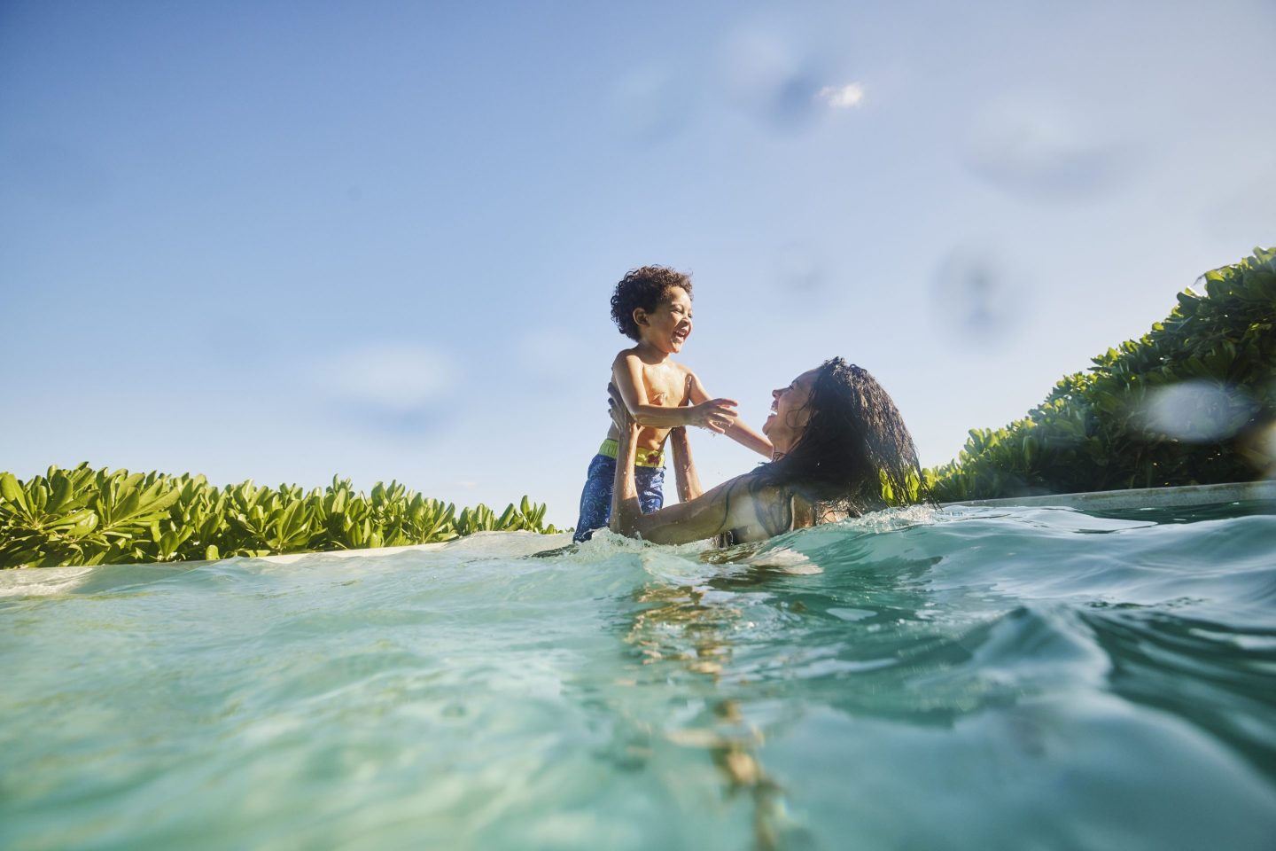 Wide shot of laughing mother and son playing in pool at tropical resort.