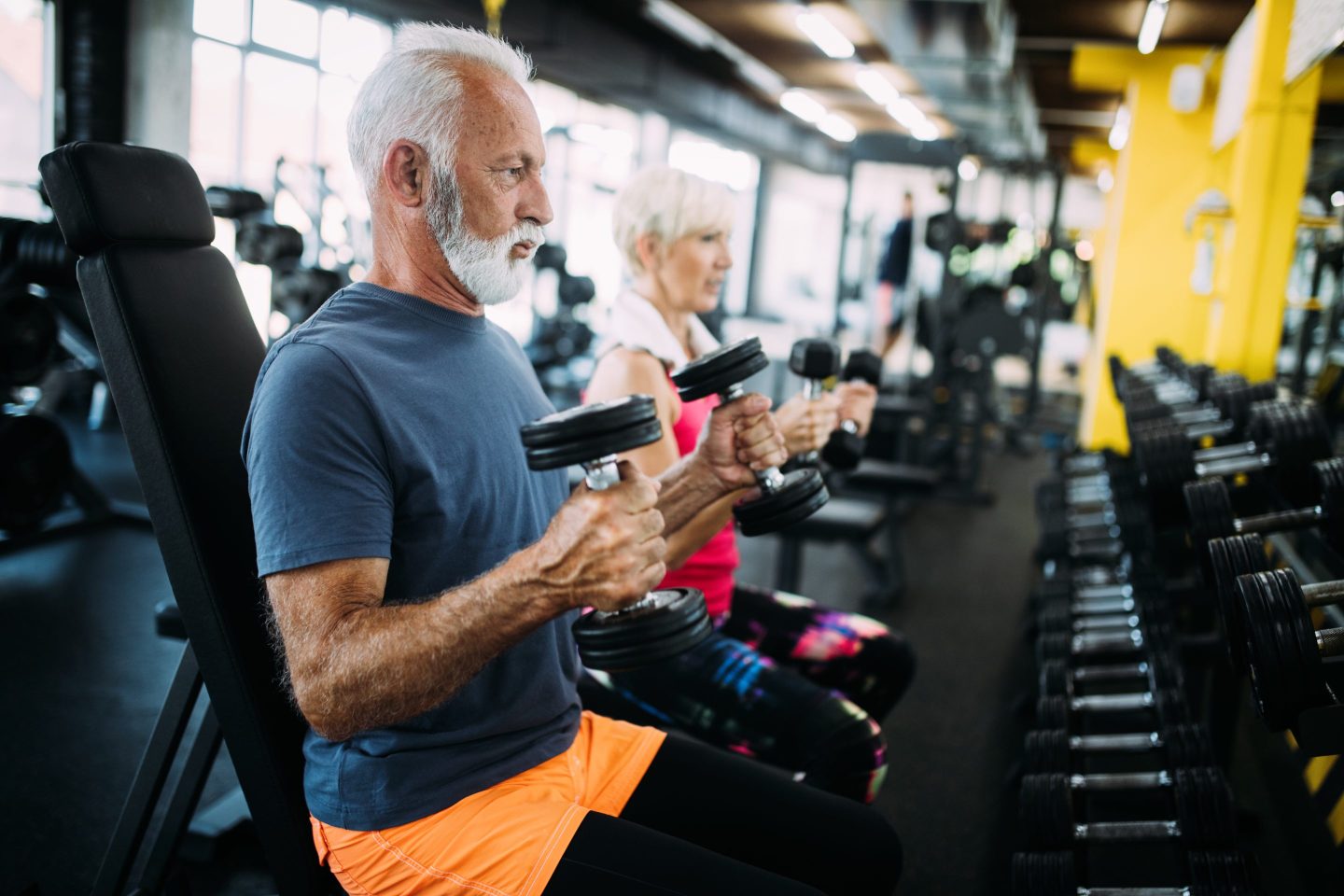 Man with gray hair and beard seated at the gym holding weights; woman with weights and gray hair in background