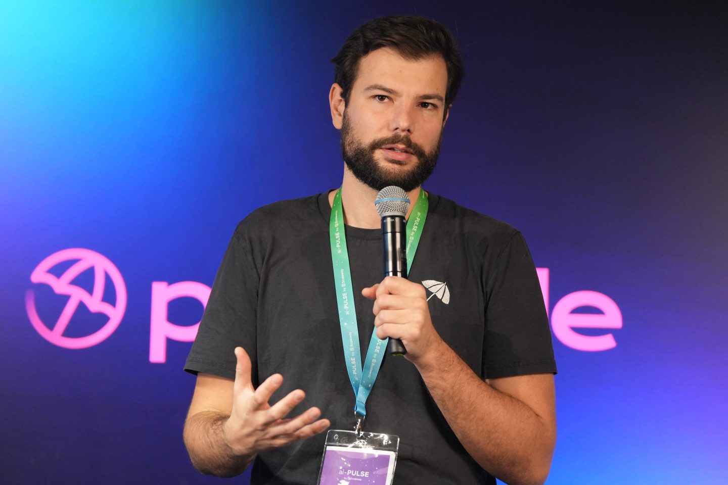 Eiso Kant, co-founder of Poolside, speaking at the ai-Pulse conference held at the Station F technology campus in Paris, France, on November 17, 2023. Wearing a black t-shirt and a conference badge, he holds a microphone while gesturing with his left hand.