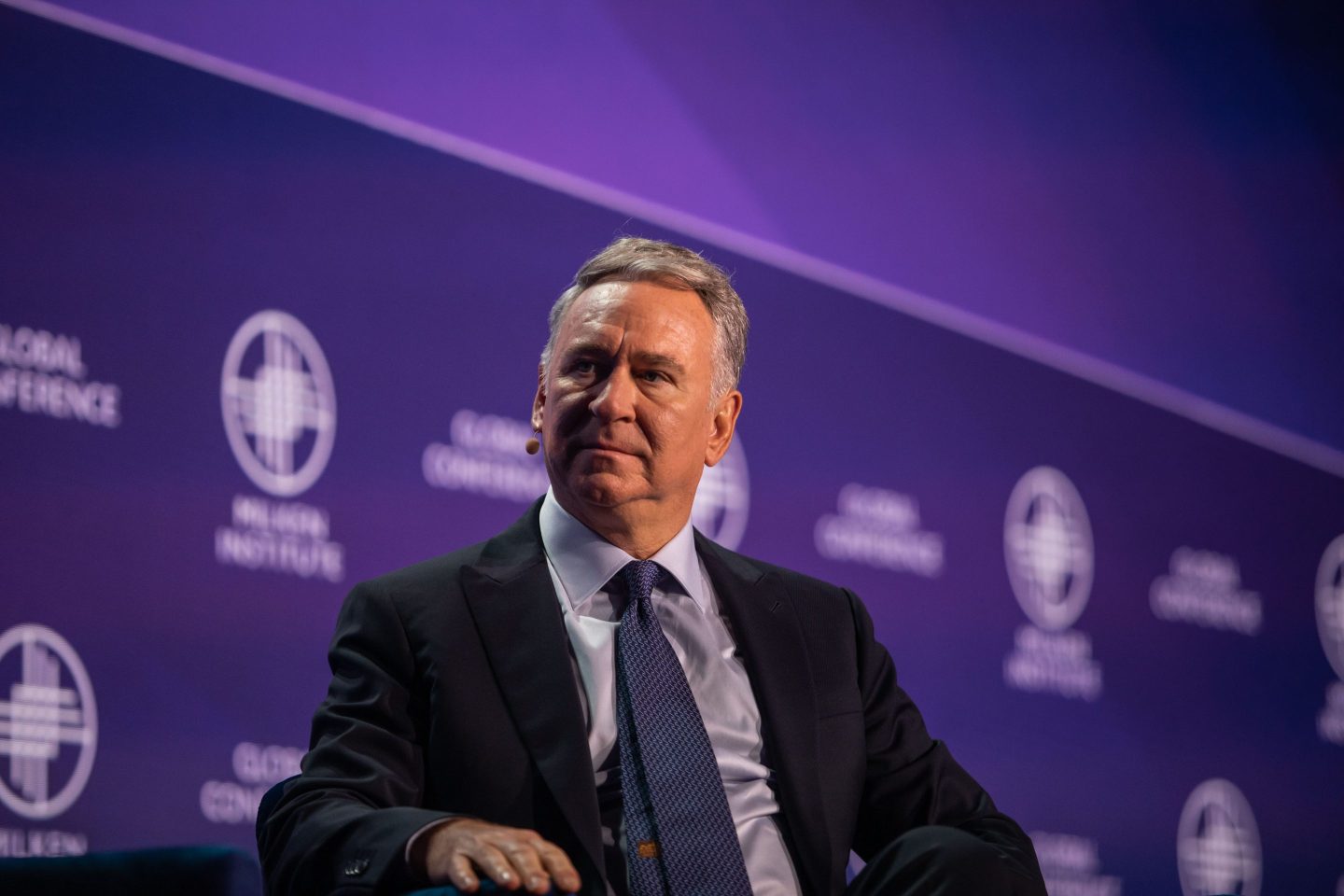 A middle-aged man, Ken Griffin, wearing a dark suit and light blue tie, sits on stage during a conference. He has short, neatly combed gray hair and is looking slightly to his left with a serious expression. Behind him is a purple backdrop with a repeating logo for the Milken Institute Global Conference.