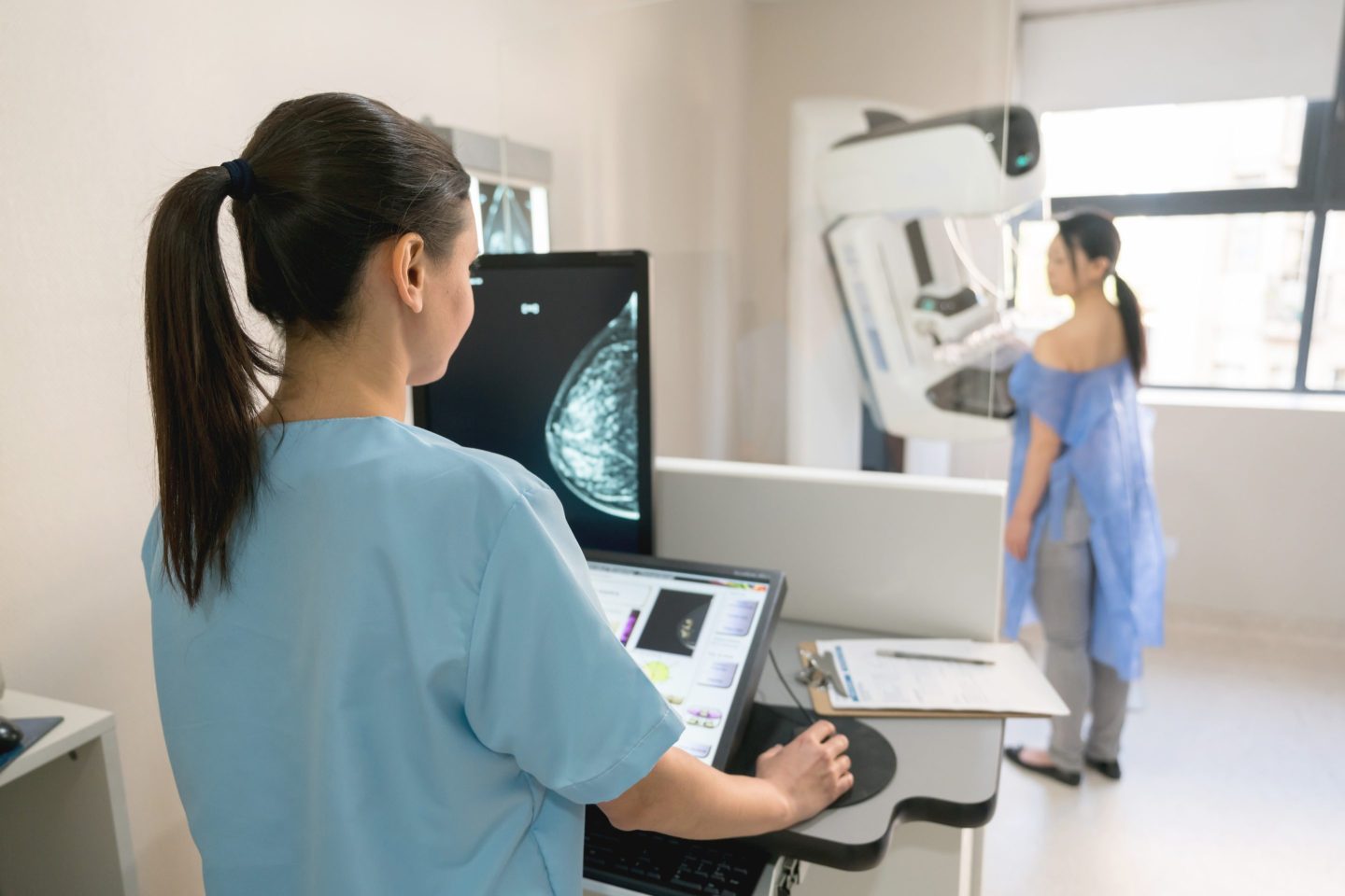 woman with her back to us looking at a radiology screen of a breast, woman in background standing at mammography machine