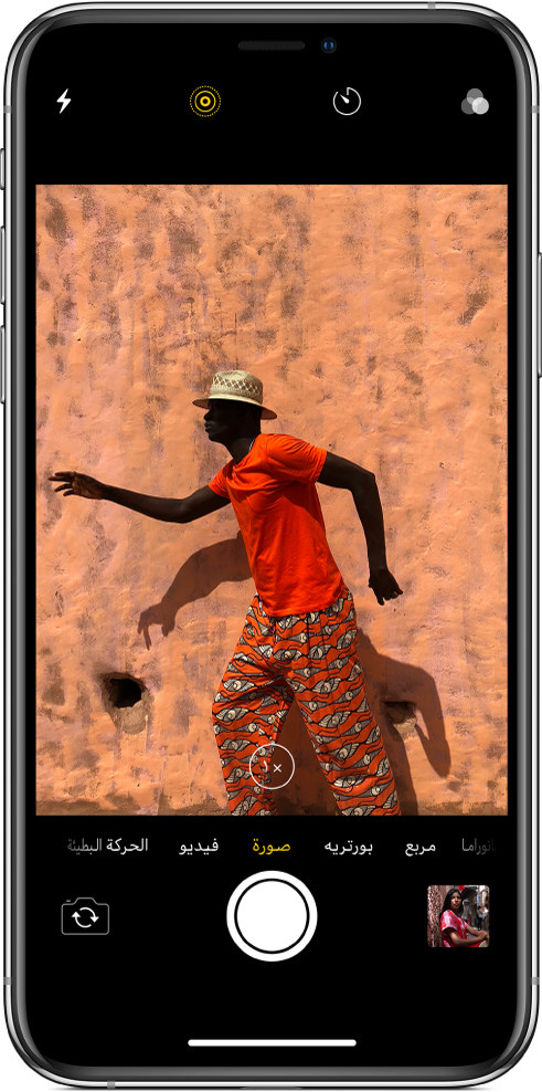 الكاميرا في نمط الصورة، مع الأنماط الأخرى على اليمين واليسار أسفل العارض. تظهر أزرار الوميض وLive Photo والمؤقت والفلاتر في أعلى الشاشة. صورة مصغرة في أسفل اليمين لتمكين الوصول إلى عرض الصور والفيديوهات الموجودة. زر الغالق في أسفل المنتصف، وزر تبديل الكاميرا في أسفل اليسار.