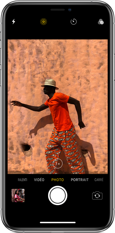 L’appareil photo en mode Photo, avec les autres modes à gauche et à droite sous le visualiseur. Les boutons pour le flash, le mode Live Photo, le retardateur et les filtres apparaissent en haut de l’écran. Touchez la vignette située en bas à gauche pour accéder aux photos et aux vidéos existantes. Le bouton de l’obturateur se trouve en bas au centre et le bouton Changer de caméra est dans le coin inférieur droit.