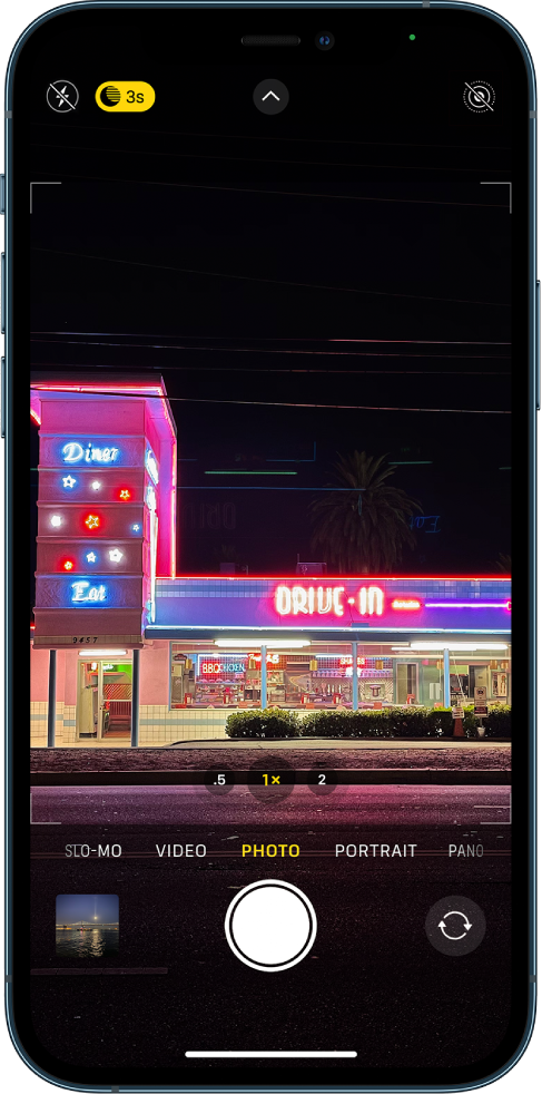 Lietotnes Camera ekrāns režīmā Photo. Zibspuldzes un nakts režīma pogas ir redzamas ekrāna augšējā kreisajā stūrī. Zibspuldze ir izslēgta, un ir aktivizēts nakts režīms. Augšā pa vidu ir poga Camera Controls; augšējā labajā stūrī ir poga Live Photo. Ekrāna apakšā no kreisās puses uz labo ir pogas Photo and Video Viewer, Take Picture un Camera Chooser Back-Facing.