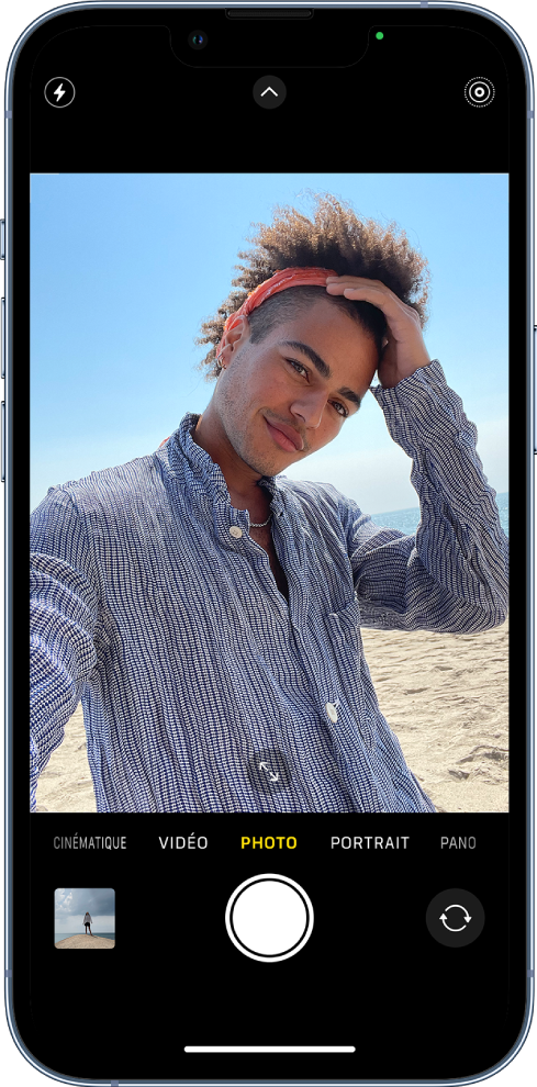 L’écran Appareil photo en mode Photo, avec les autres modes à gauche et à droite sous le viseur. En dessous des différents modes d’appareil photo, de gauche à droite, se trouvent le bouton « Visualiseur de photos et vidéos », le bouton « Prendre une photo » et le bouton « Sélecteur de caméra - face arrière ». Les boutons pour le flash, les commandes de l’appareil photo et le mode Live Photo figurent en haut de l’écran. Dans le cadre de l’appareil photo, le sujet prend un selfie.