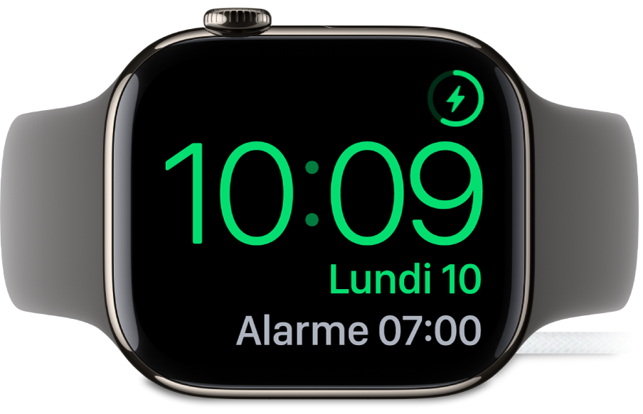Apple Watch placée sur sa tranche et connectée au chargeur. L’écran montre le symbole de recharge dans le coin supérieur droit, l’heure actuelle en dessous et l’heure de la prochaine alarme.
