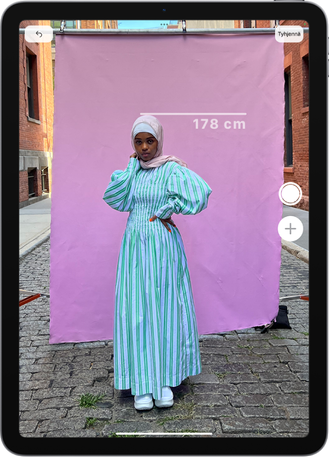 Henkilön pituus mitataan Mittanauha-apissa, ja lukema tulee näkyviin henkilön pään yläpuolelle. Ota kuva -painike on aktiivinen oikeassa alakulmassa, jotta voit ottaa kuvan mittauksesta. Vihreä kameran käytön merkki näkyy ylhäällä.