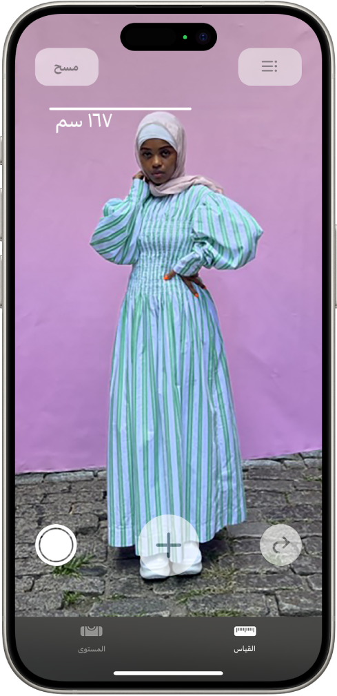 شخص يتم قياس طوله في تطبيق القياس، ويظهر قياس الطول أعلى رأس الشخص. زر التقاط صورة نشط بالقرب من الزاوية السفلية اليسرى لالتقاط صورة للقياس. ويظهر مؤشر الكاميرا المستخدمة الأخضر في الأعلى.