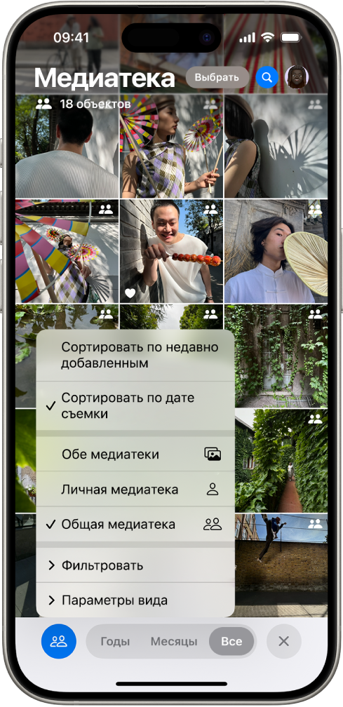 Общая медиатека iCloud в приложении «Фото». Выбрана кнопка «Общая медиатека» в левом нижнем углу, и в списке вариантов выбрана общая медиатека. В правом верхнем углу всех отображающихся в медиатеке фотографий стоит значок общей медиатеки.