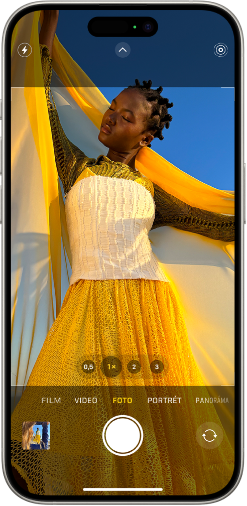 Kamera v režime Foto s ďalšími režimami zobrazenými vľavo a vpravo pod hľadáčikom. V hornej časti obrazovky sa nachádzajú tlačidlá Blesk, Ovládanie kamery a Live Photos. V ľavom dolnom rohu sa nachádza tlačidlo prehliadača fotiek a videí. Dole uprostred sa nachádza tlačidlo Odfotiť a vpravo dole tlačidlo na výber kamery prepnuté na zadnú kameru.