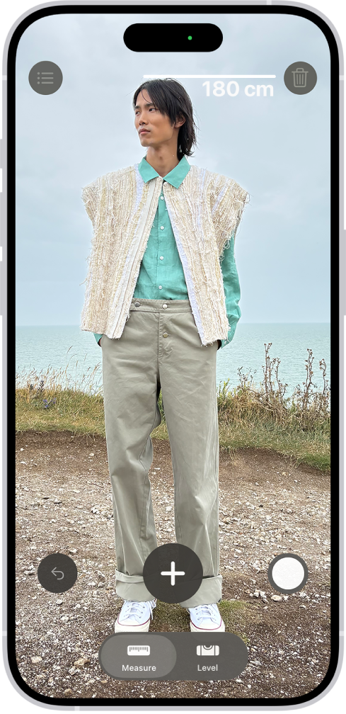 Iemands lengte wordt gemeten met de Meten-app, waarbij de gemeten lengte boven het hoofd van de persoon wordt weergegeven. Met de knop 'Maak foto' rechtsonderin kan een foto van de meting worden gemaakt. De groene indicator die aangeeft dat de camera in gebruik is, staat bovenin.